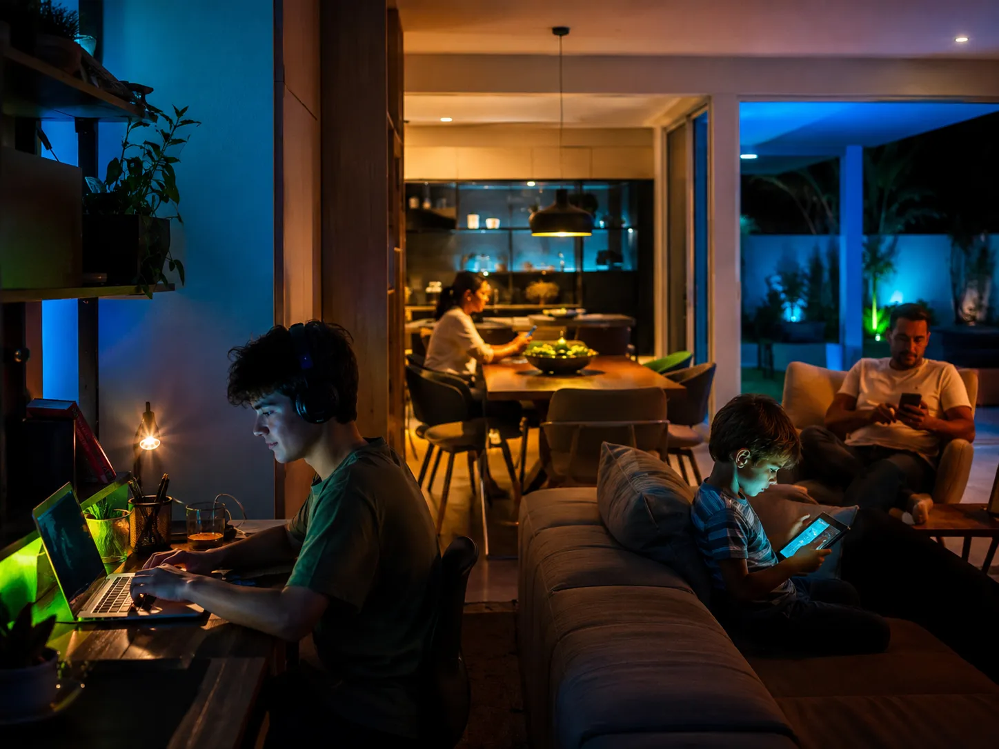 Família em casa à noite, cada um usando um dispositivo conectado ao mesmo tempo — notebook, tablet e celulares.